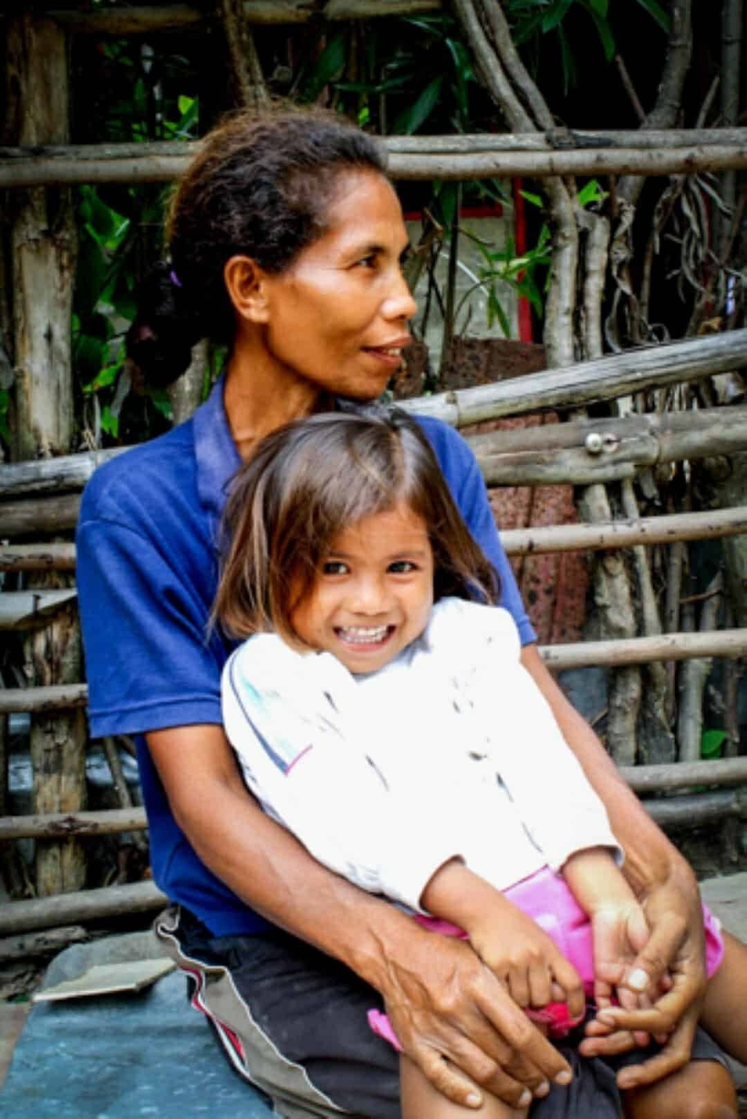 Mother holds smiling young girl securely on her lap, arms wrapped around her, outdoors by a wooden fence