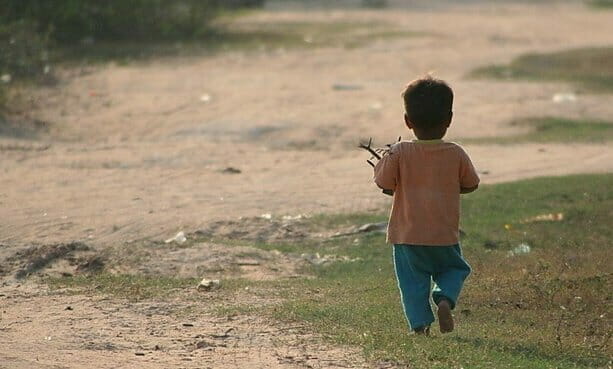 A young child walks alone barefoot along a dirt path, seen from behind