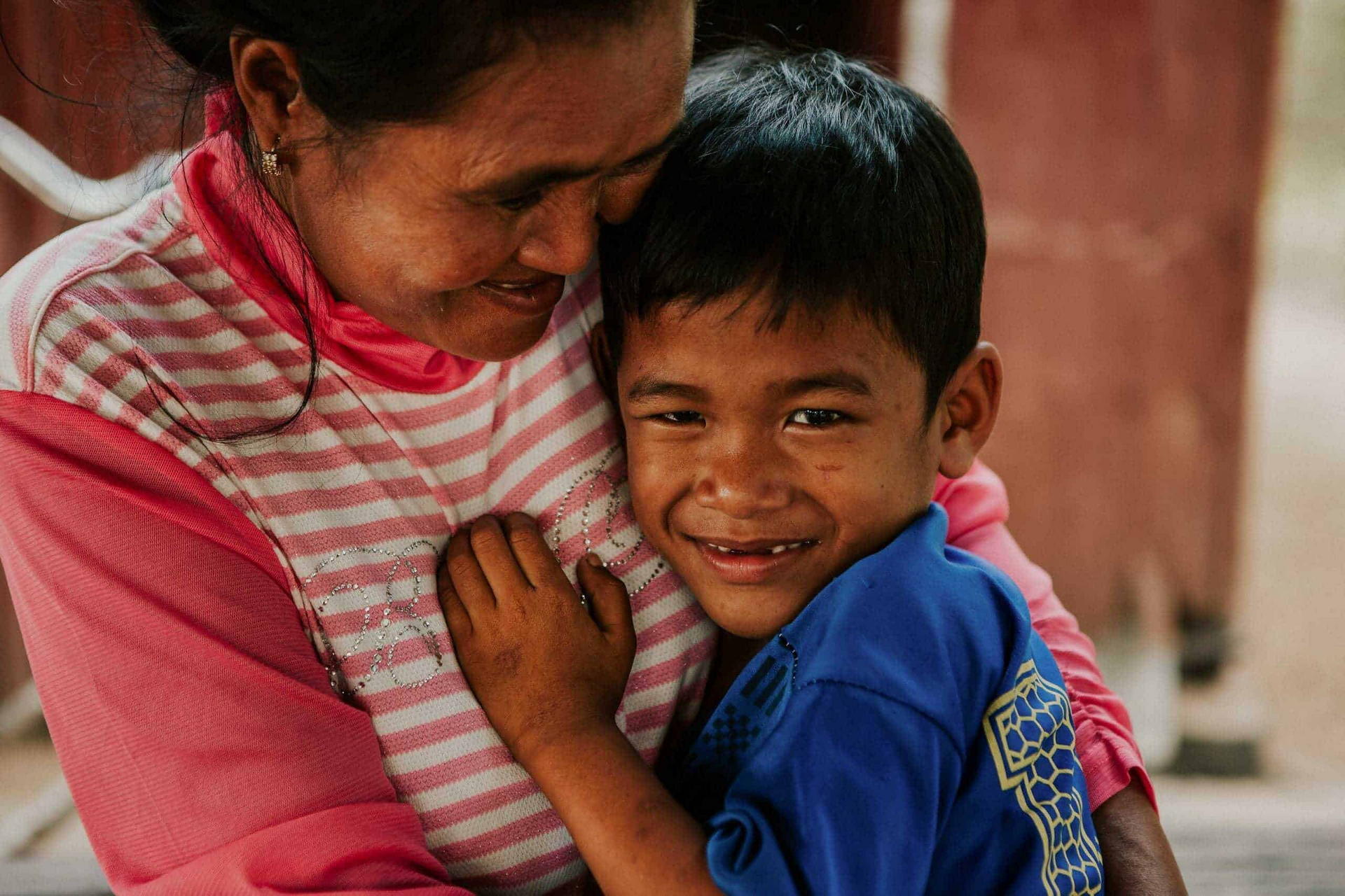 A woman embraces a smiling young boy warmly, holding him close as he looks toward the camera