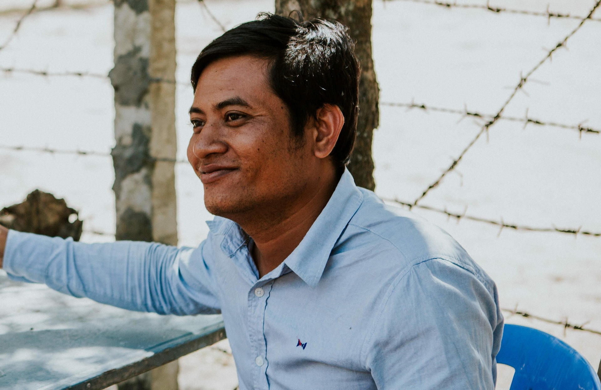Leak sits at a table outdoors, smiling, with a barbed-wire fence visible behind him