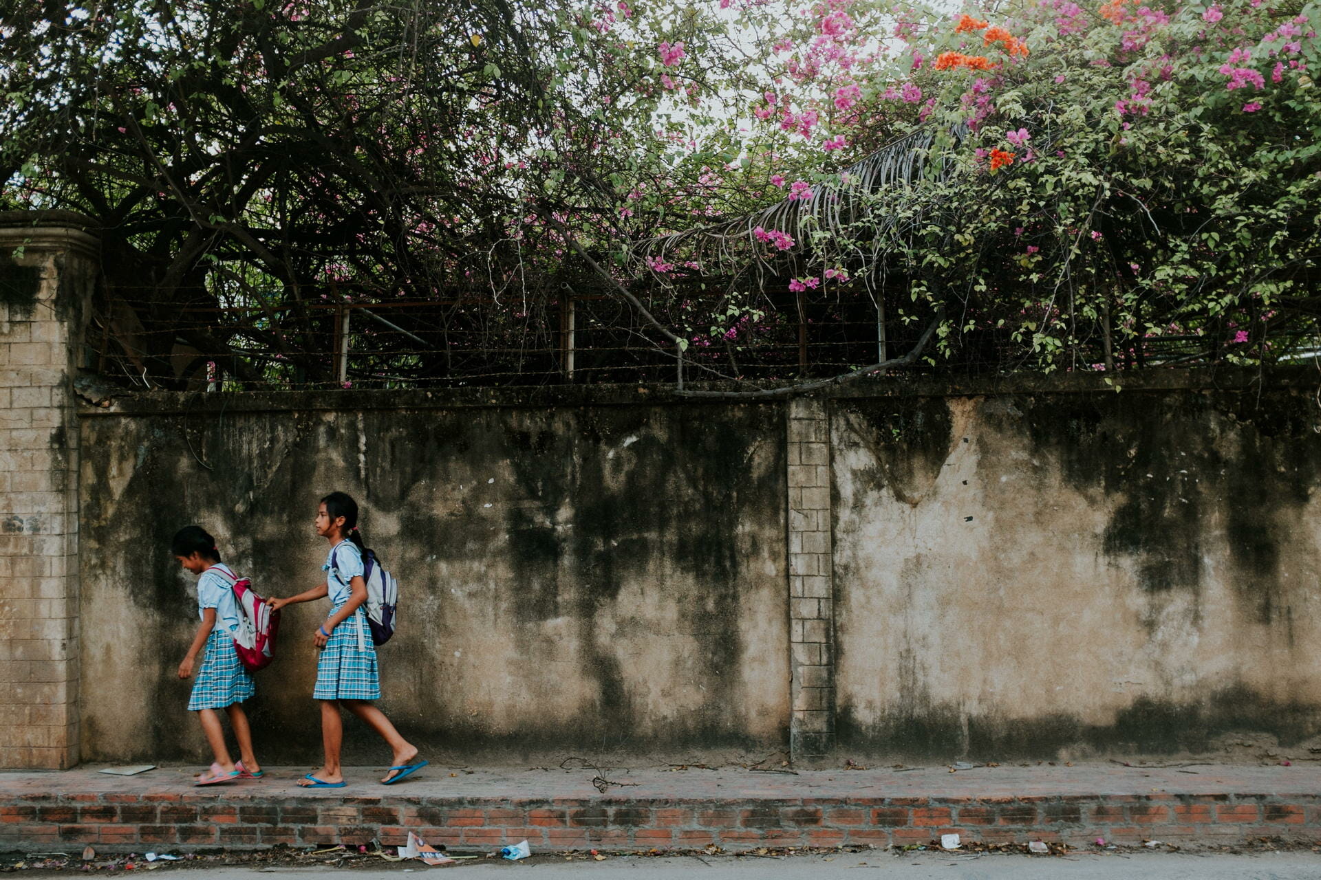 Two girls in matching school uniforms and backpacks walk along a sidewalk beside a weathered wall with flowering vines above