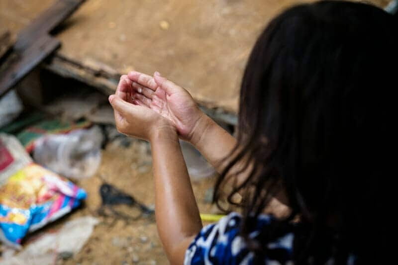 A child in Cambodia sits with cupped hands raised, in a simple dirt-floored home setting