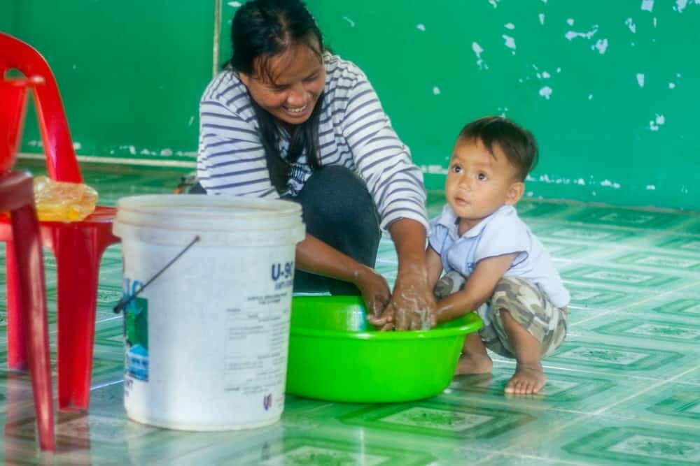 A mother smiles while helping her young son wash his hands together in a green basin