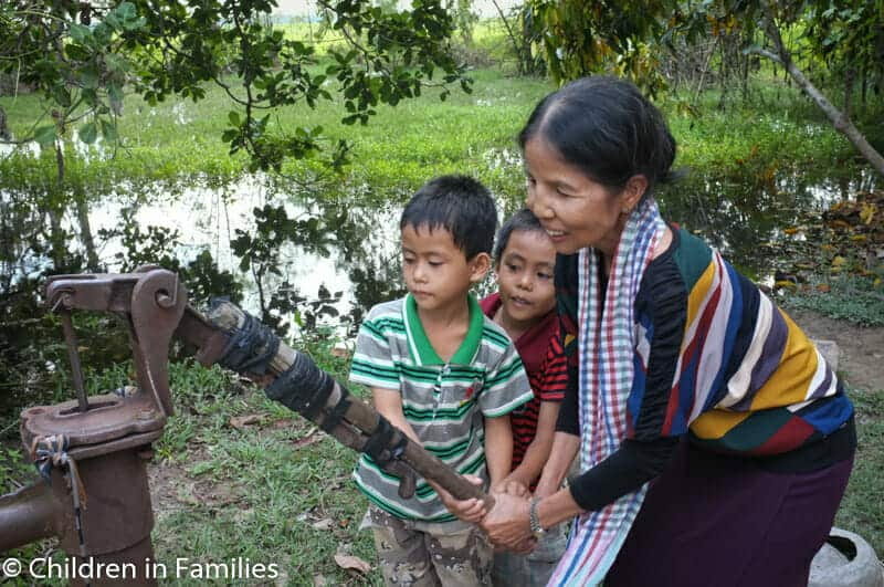 An older woman helps two young boys pump water from a well near a green, flooded field