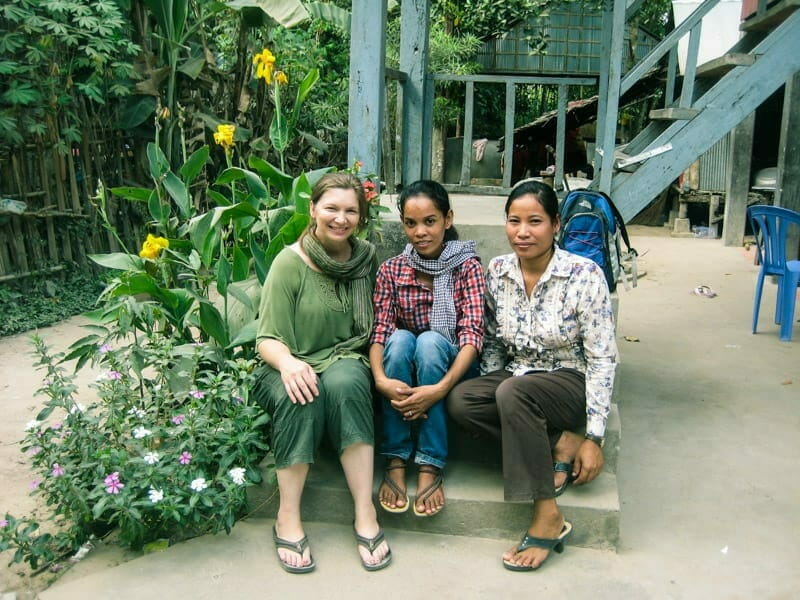 Three women sit together smiling on steps outside a stilted house, surrounded by garden flowers
