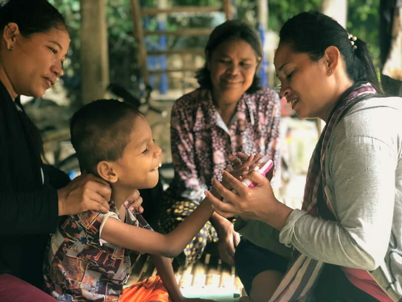 A smiling child reaches toward a woman's hands while other women look on warmly in a village setting