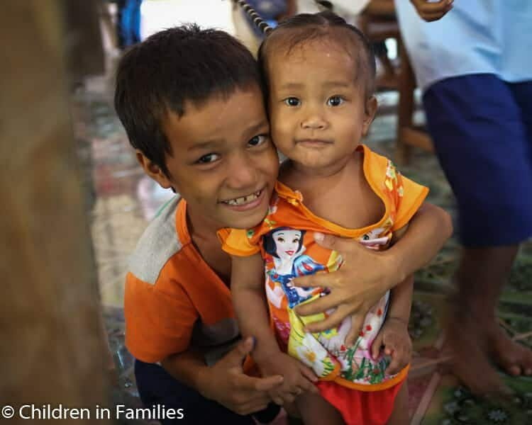 A young boy smiles while affectionately holding a toddler girl in his arms indoors