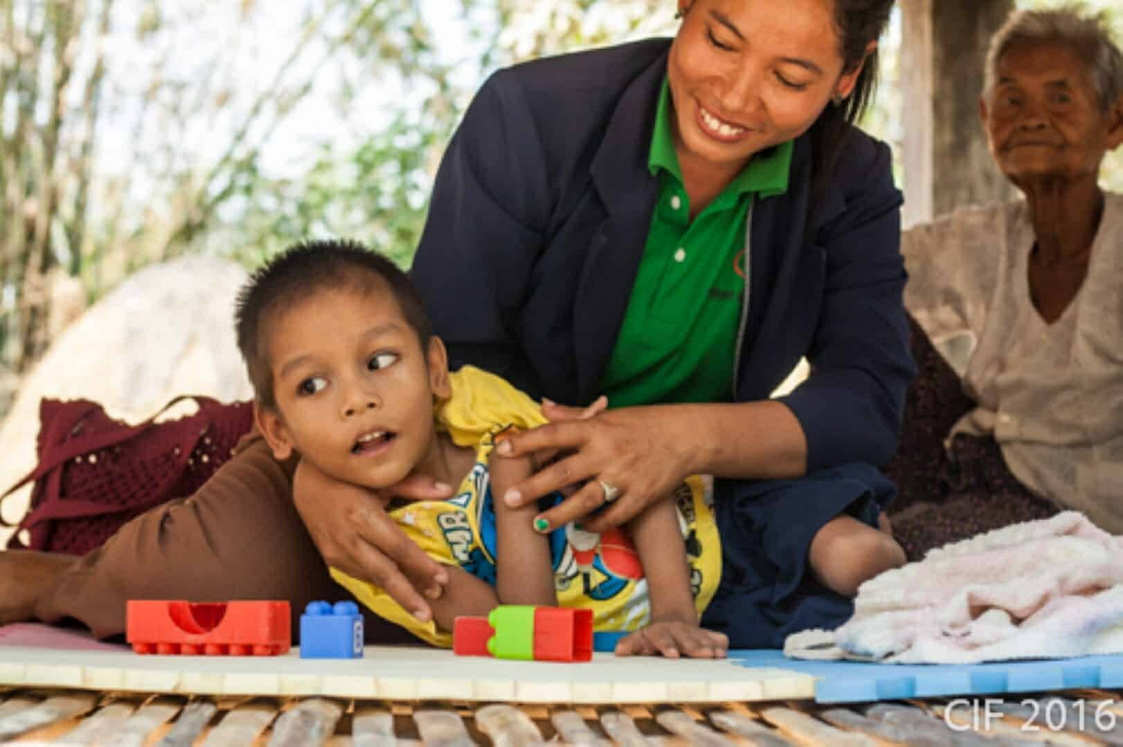 A worker gently supports a young boy with a disability as he plays with blocks during a home visit, with his grandmother nearby
