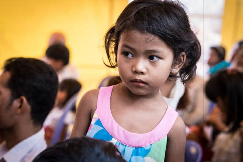 Young girl in a colorful dress looking thoughtfully to the side amid a seated crowd at a gathering