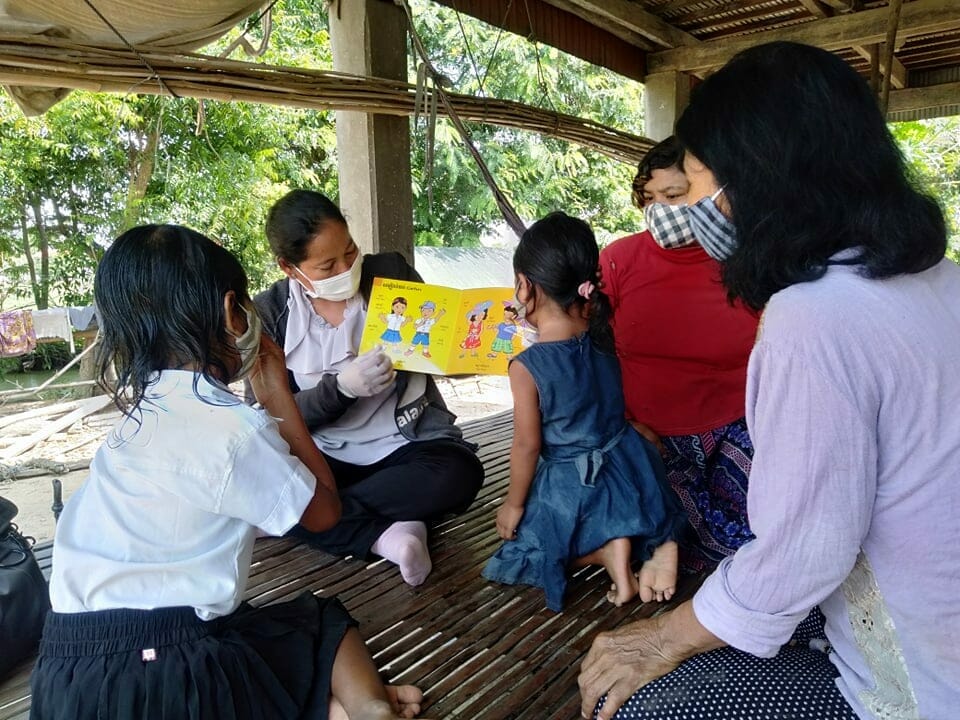 A masked worker reads a picture book to children and family members gathered on a wooden platform outdoors