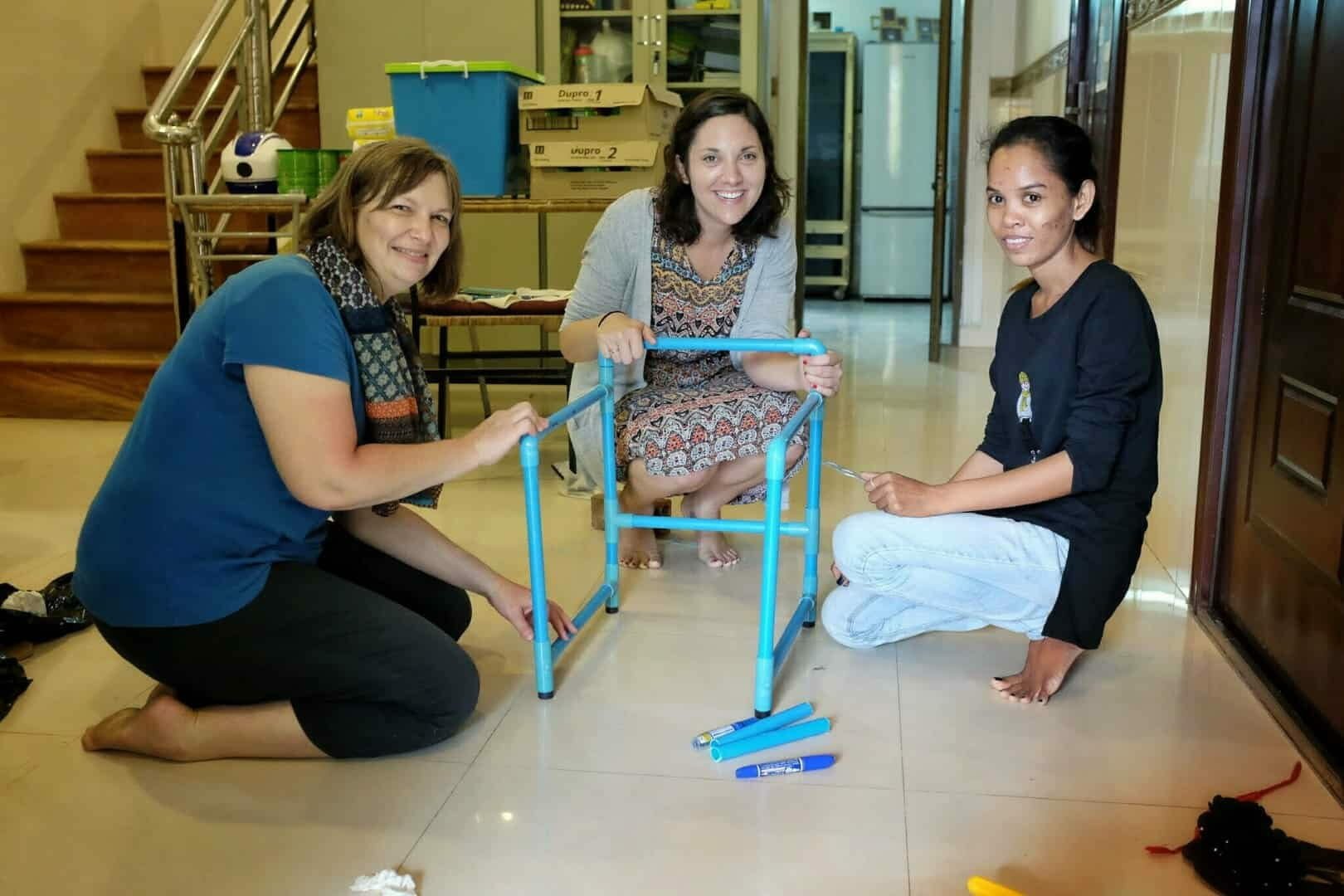 Three women kneel together assembling a blue PVC pipe walking frame on a tiled floor
