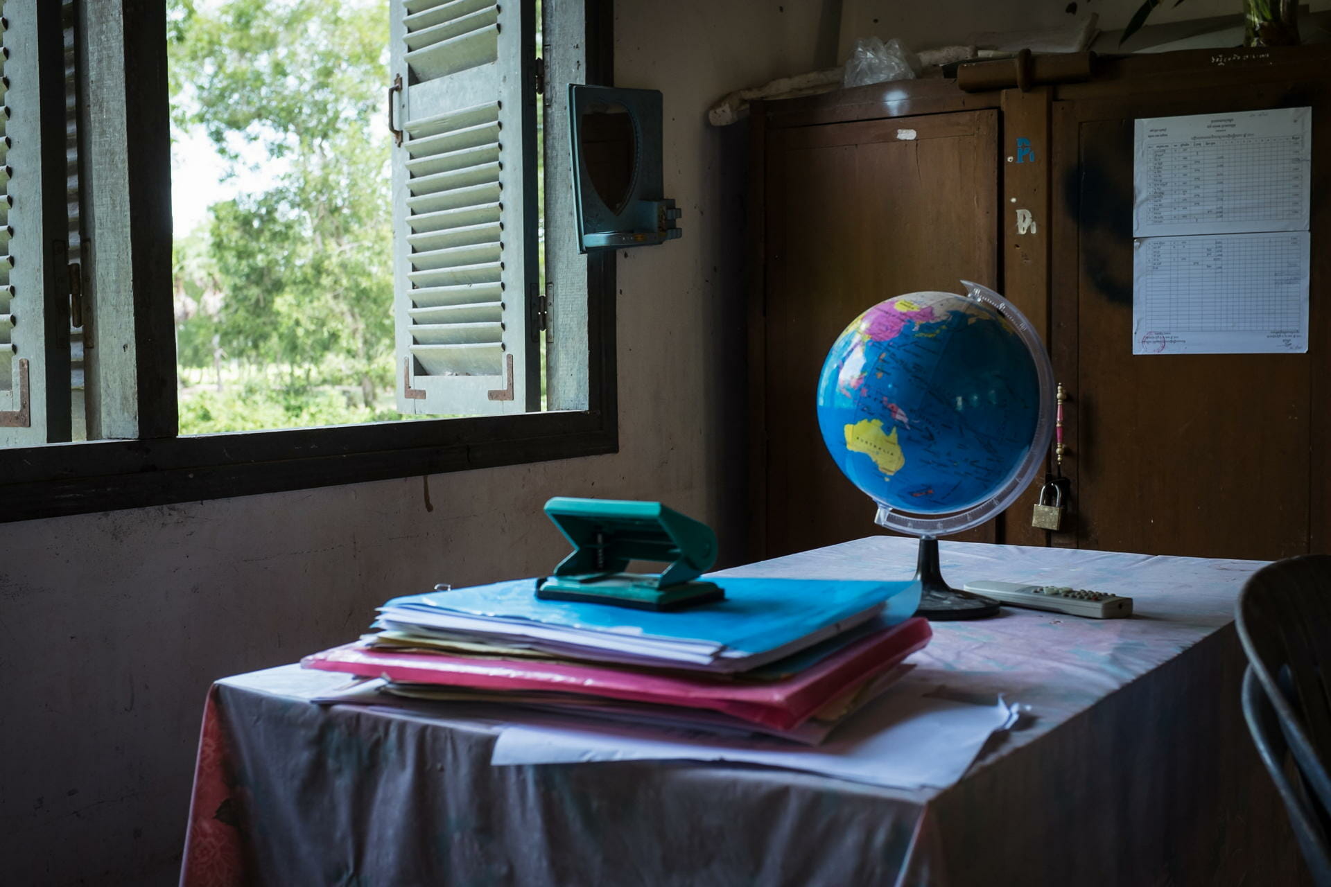A desk with a globe, folders, and a hole punch inside a modest schoolroom, sunlight through wooden shutters