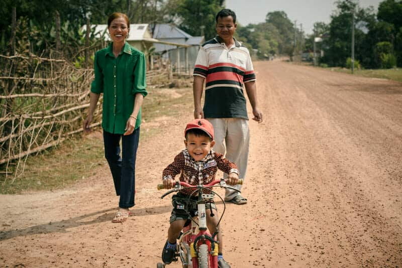 Foster parents smile as their young son rides a bicycle ahead of them on a dirt road