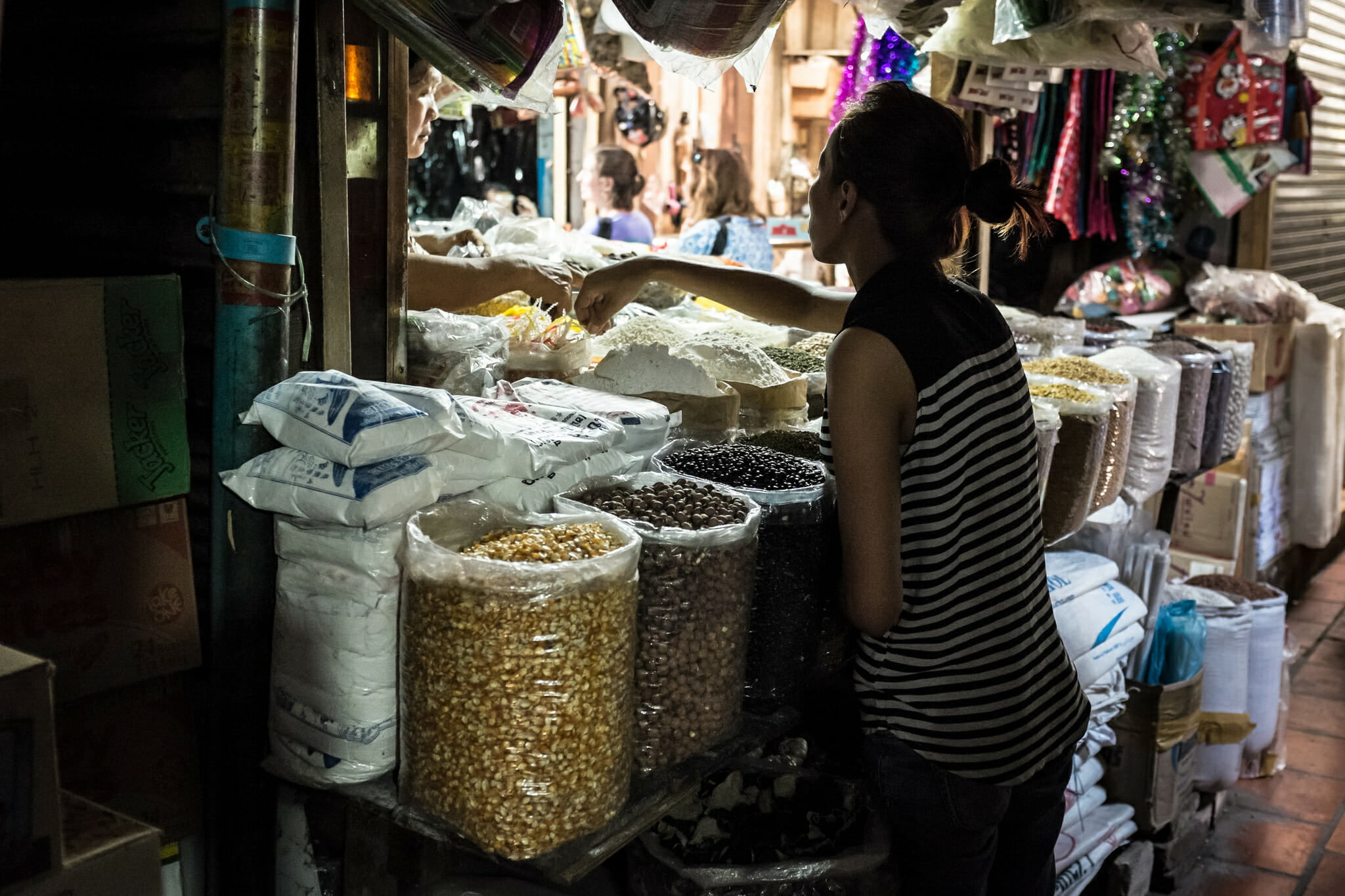 A woman shops at a market stall filled with sacks of grains, beans, and rice, exchanging goods with a vendor