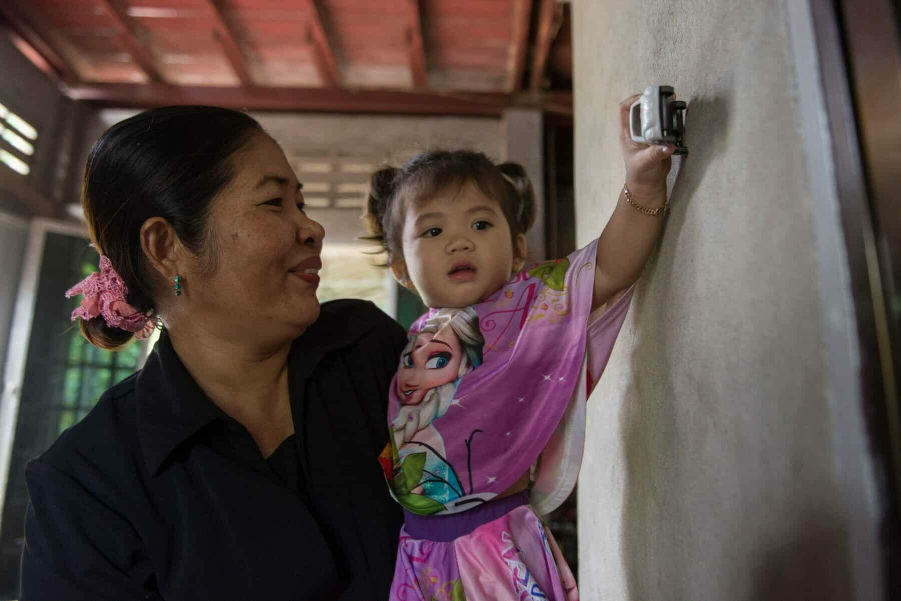 Chanthy holds a young child who reaches toward the wall with a toy car, both smiling warmly at home