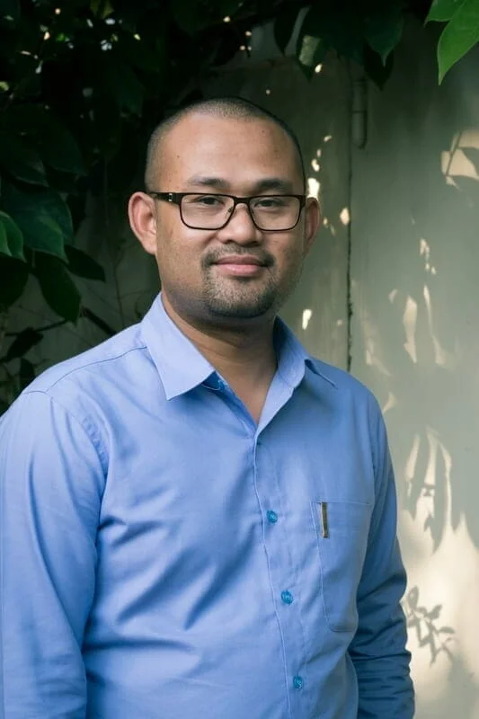 Dy Noeut smiling in a blue collared shirt, standing outdoors near a leafy plant and wall