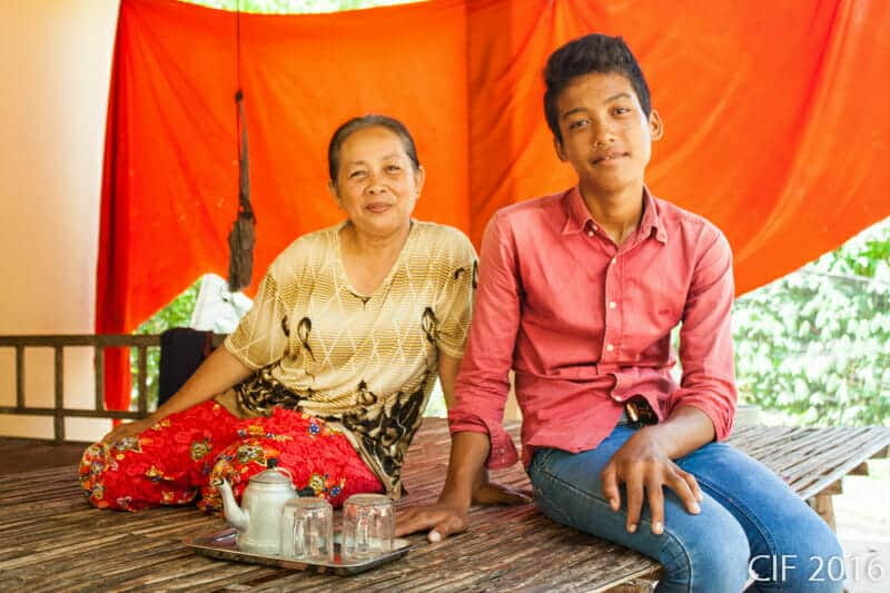 A foster mother and her teenage son sit together on a bamboo platform, smiling for the camera
