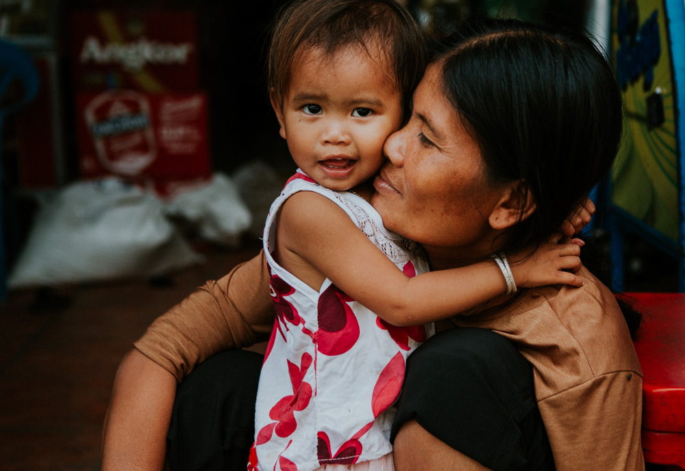A mother holds and kisses her young daughter, who smiles as she clings to her shoulder