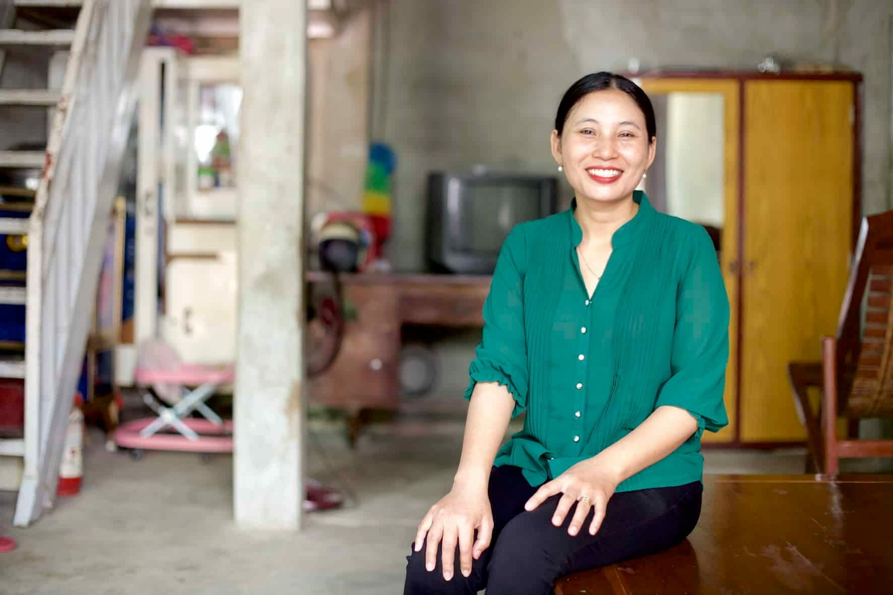 Woman in a green blouse smiles while sitting in her home, with children's toys visible in the background