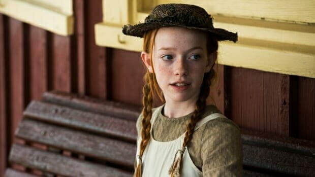 Red-haired girl with braids and straw hat, dressed as Anne Shirley, standing by a train station