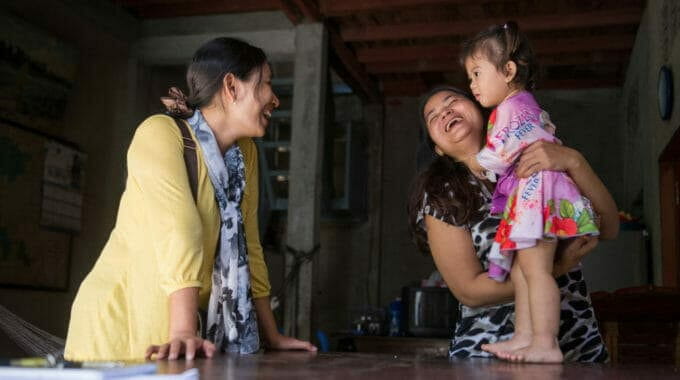Two women share a joyful laugh as one holds a smiling toddler in a floral dress