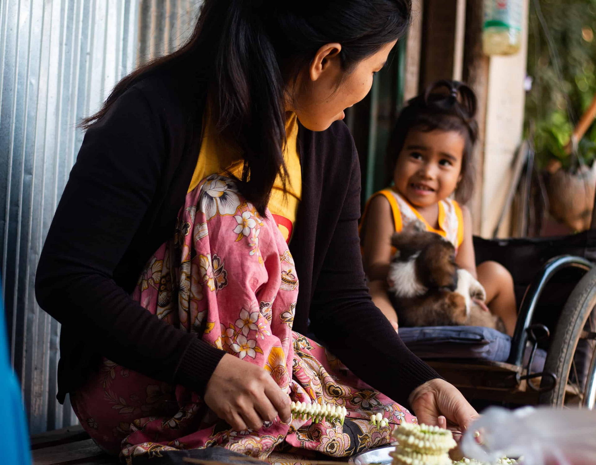 A woman threads flower garlands at home while a smiling girl nearby holds a small dog