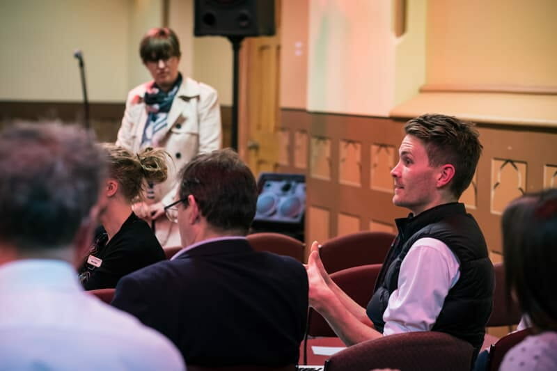 A man speaks to an audience during a discussion event, with attendees seated and listening in a hall