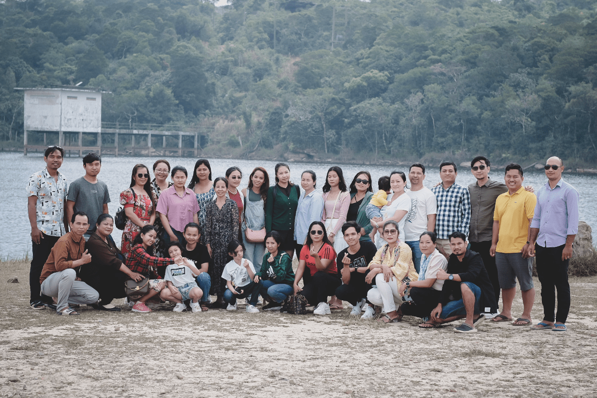 Large group of team members posing together on a sandy shore beside a river, with forested hills behind them