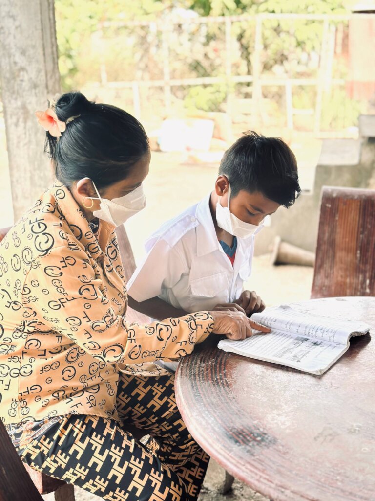 A masked woman helps a young Cambodian boy in a school uniform read from a book at an outdoor table