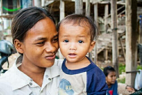 Woman smiling and holding a young child close, standing outside near wooden structures